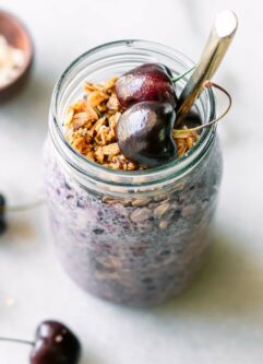 a jar of purple colored overnight oats garnished with fresh cherries on a white table