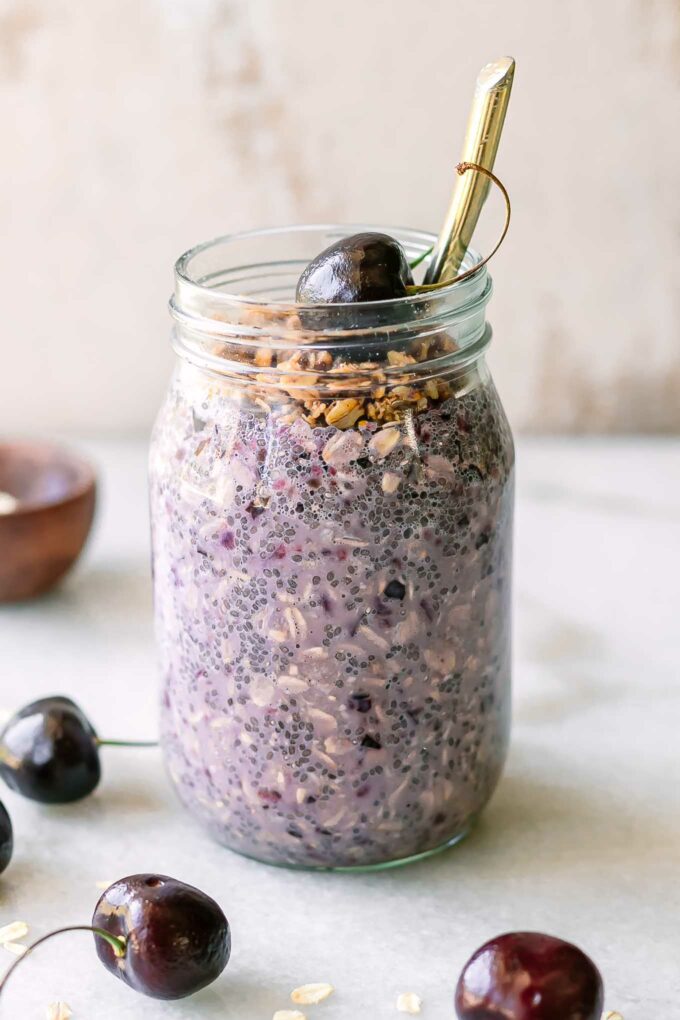 a glass jar with cherry overnight oats garnished with granola and cherries, with a gold spoon