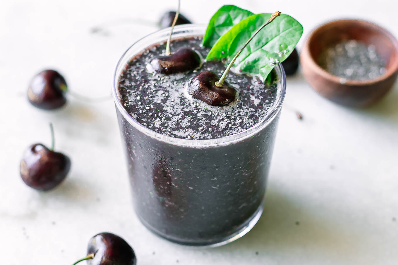 a dark purple cherry spinach smoothie in a glass with a cherry and spinach as garnish on a white table with fresh cherries