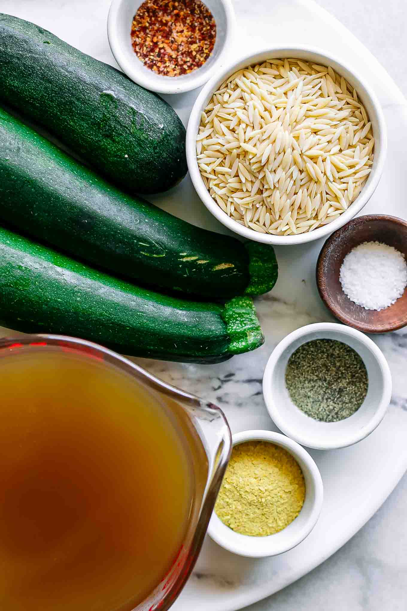 zucchini, orzo pasta, vegetable broth, and seasonings on a white table for orzotto pasta