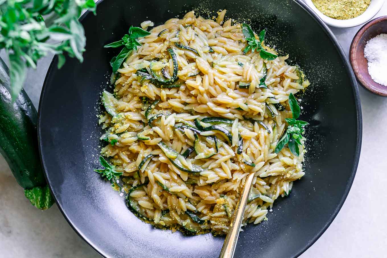 a black bowl with creamy zucchini orzotto pasta with a gold fork on a table with fresh zucchini