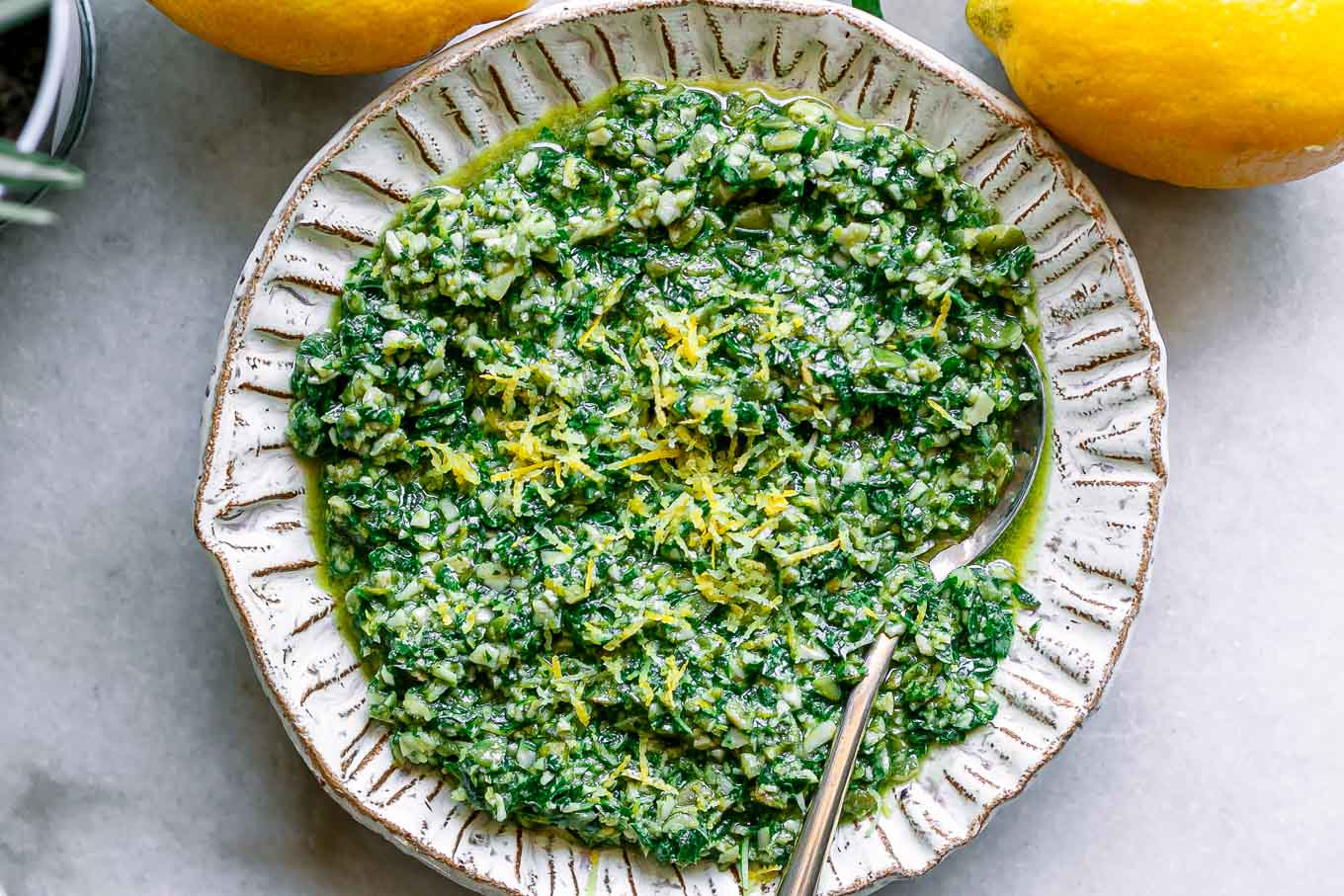 lemon pesto sauce with lemon zest as garnish in a white bowl with a gold fork on a white table