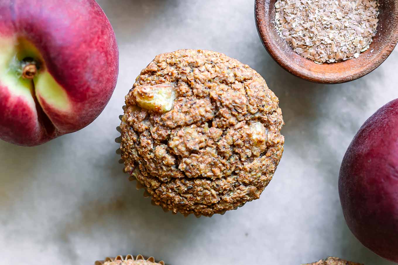 peach bran muffins on a white countertop with peaches and a bowl of wheat bran