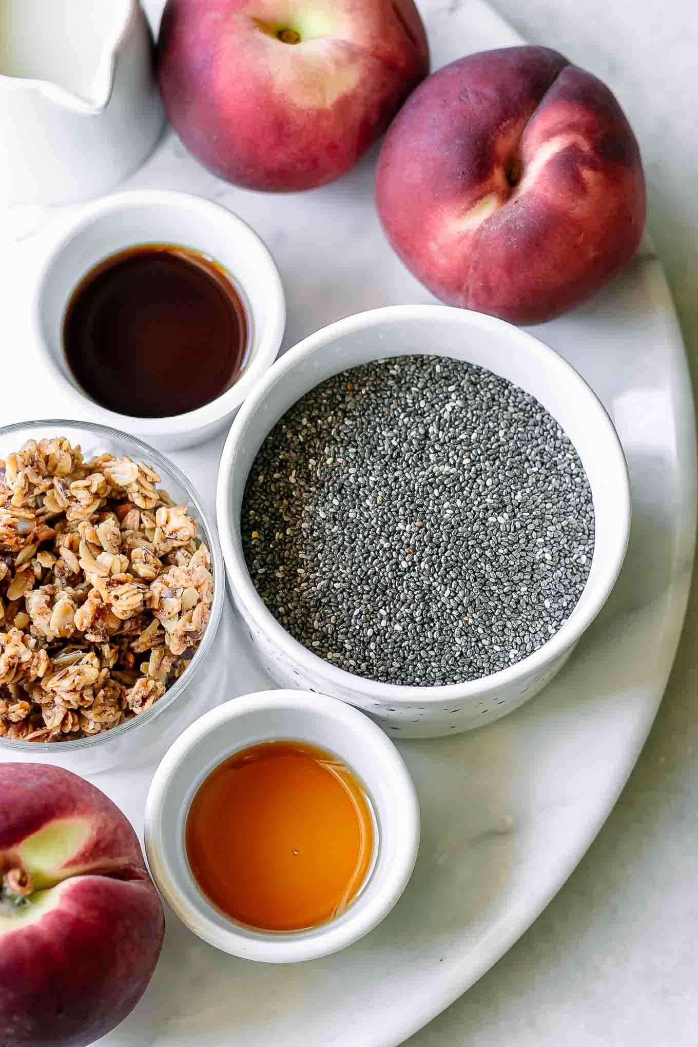bowls of chia seeds, milk, peaches, vanilla, and maple syrup for chia pudding on a white table