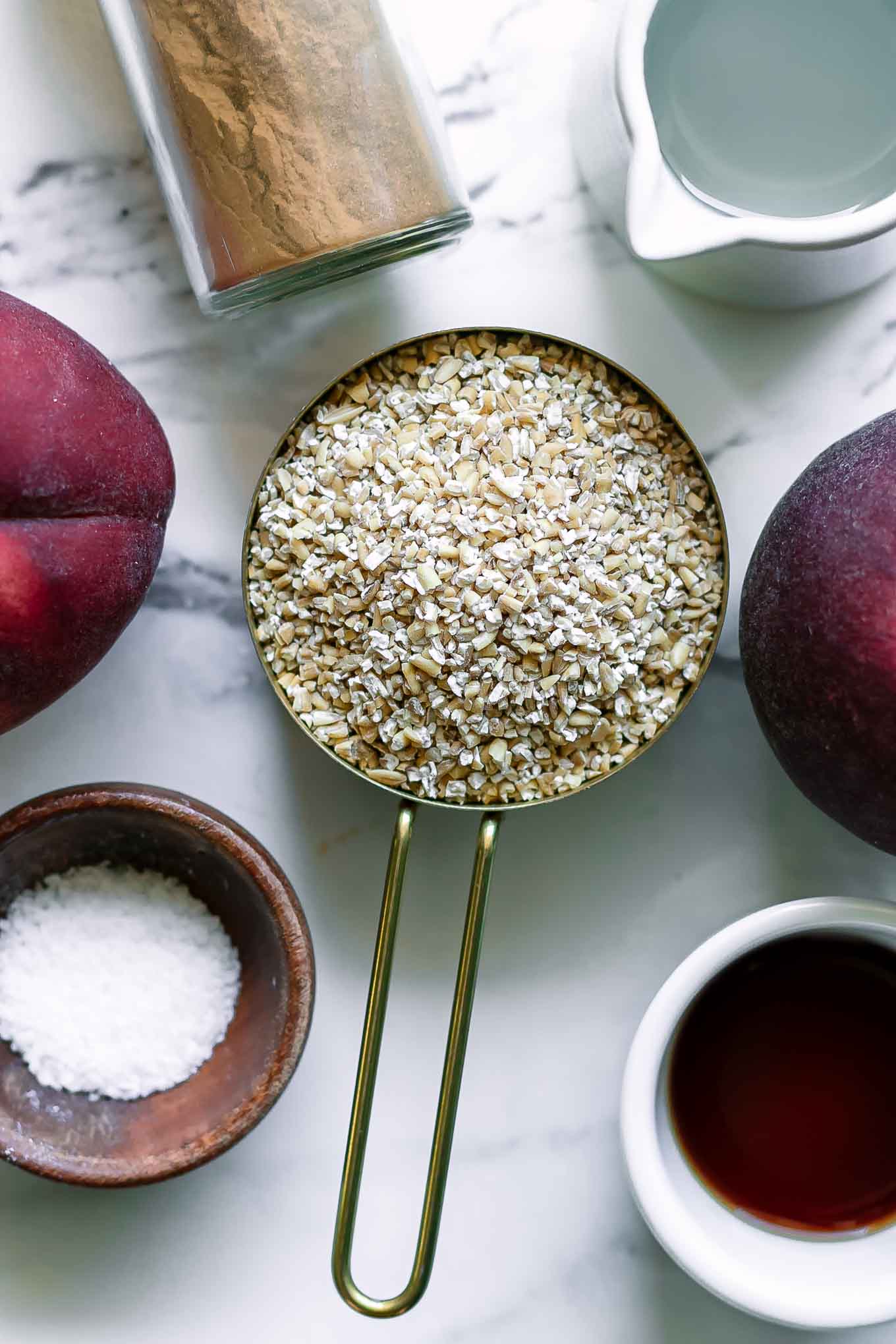 bowls of oats, cinnamon, vanilla, maple syrup, cinnamon, and whole peaches on a white table