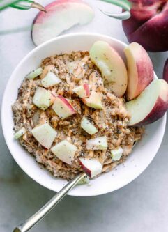 a bowl of peach oatmeal garnished with chopped peaches and nut butter on a white table with whole peaches