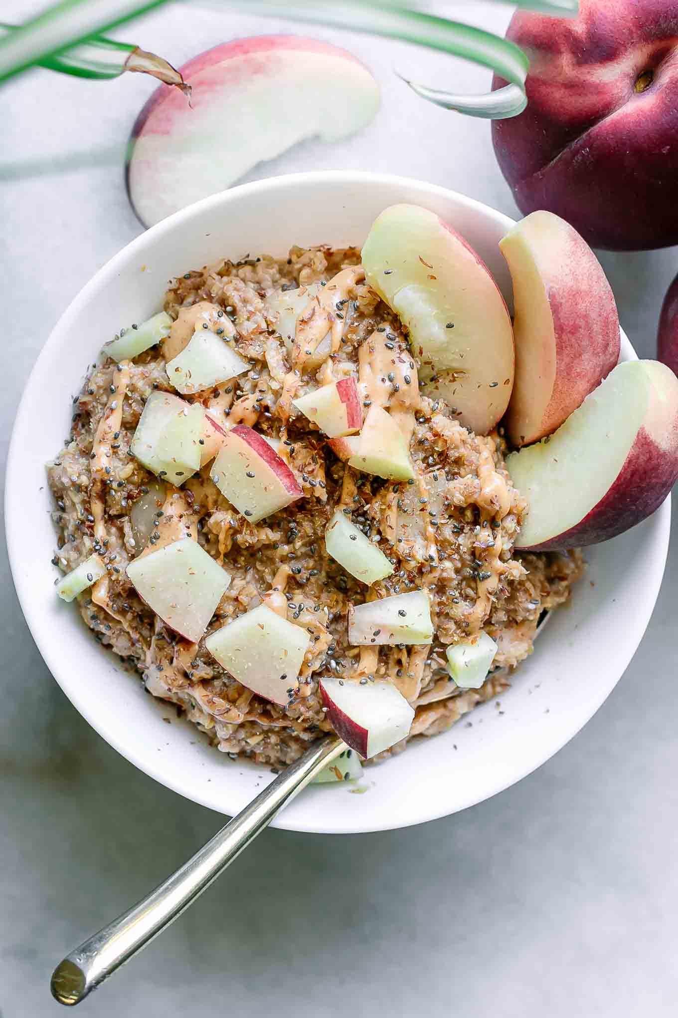 a bowl of peach oatmeal garnished with chopped peaches and nut butter on a white table with whole peaches