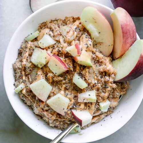a bowl of peach oatmeal with chopped peaches and drizzled nut butter on top on a table with fresh peaches