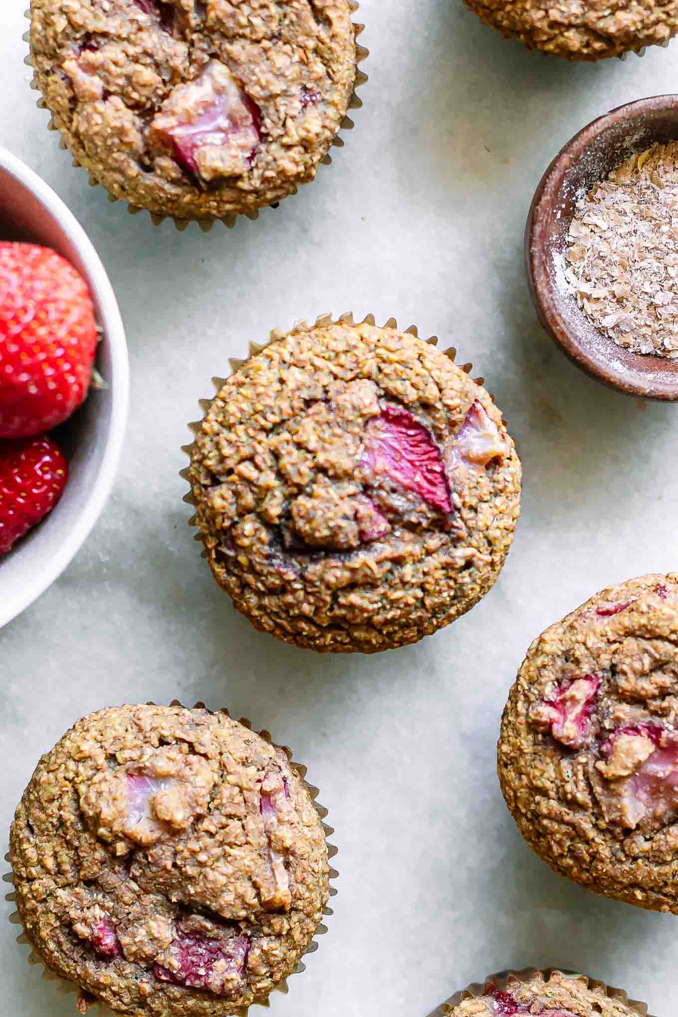 strawberry bran muffins on a white table with a bowl of strawberries and a bowl of wheat bran