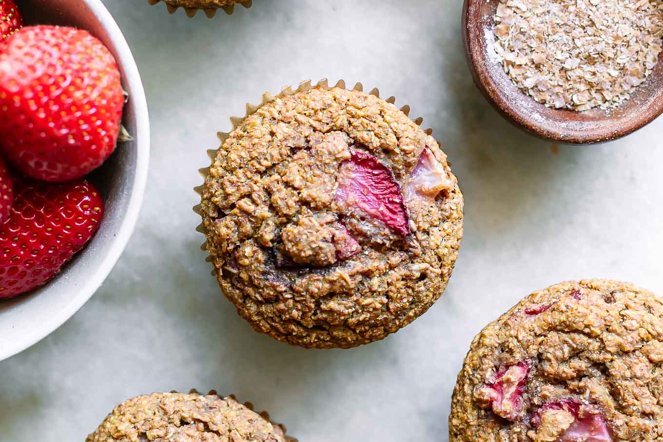 a strawberry bran muffin on a white table with a bowl of fresh strawberries