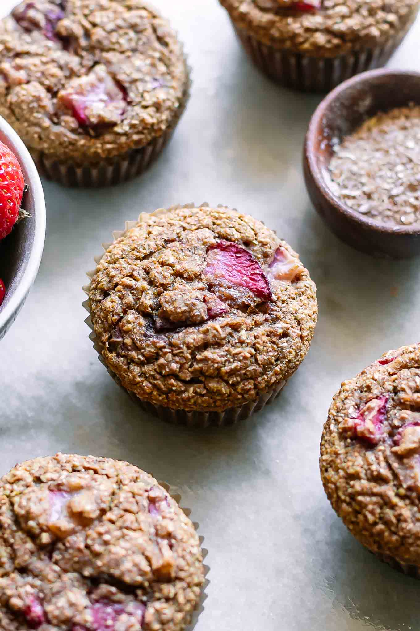 strawberry bran muffins on a white table with a bowl of strawberries and a bowl of wheat bran