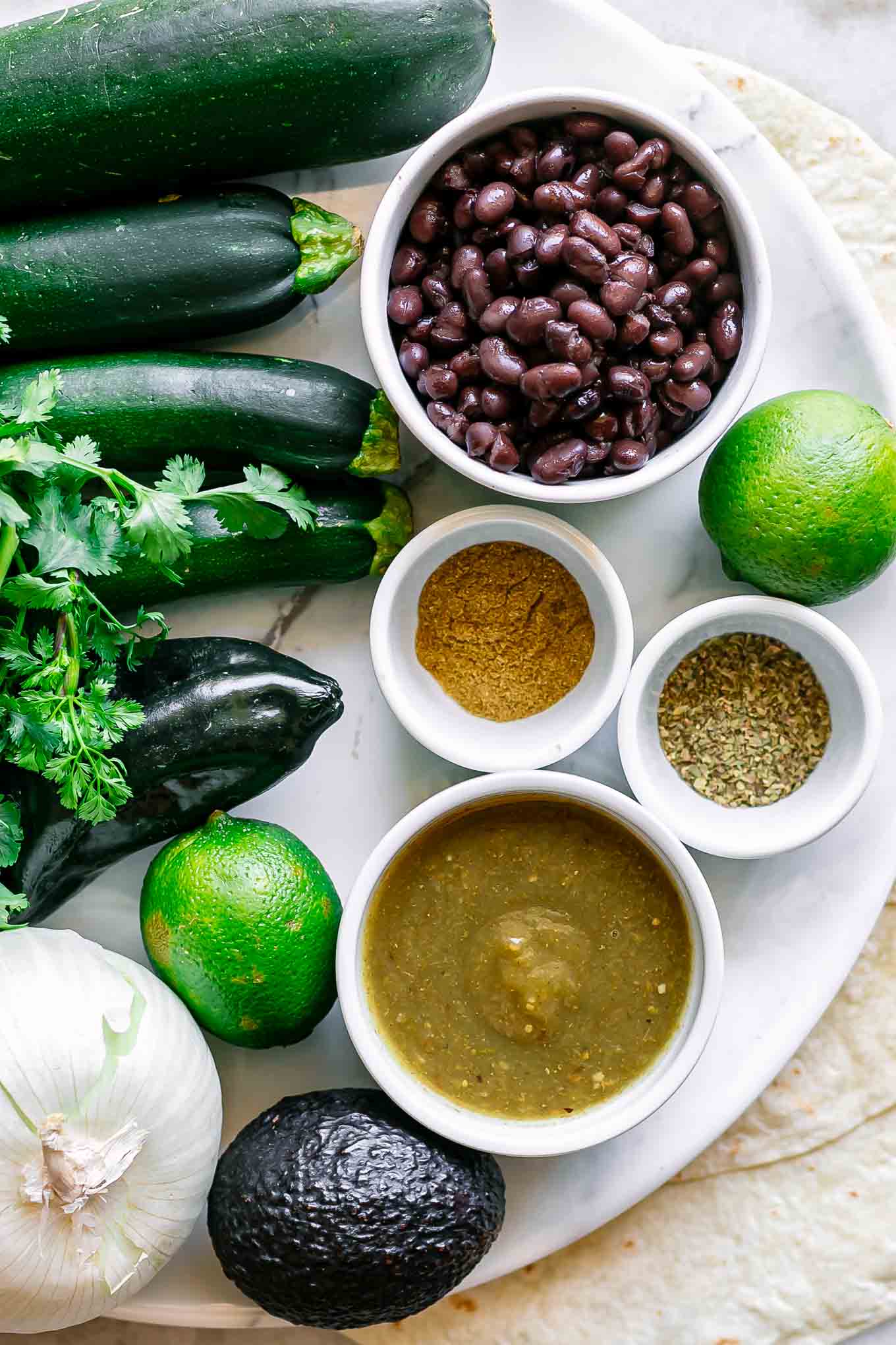 zucchini, onions, and bowls of black beans, cilantro, enchilada sauce, and spices on a table with tortillas for enchiladas