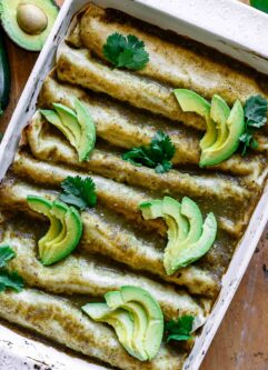 a white dish with zucchini enchiladas with green sauce topped with avocados and cilantro