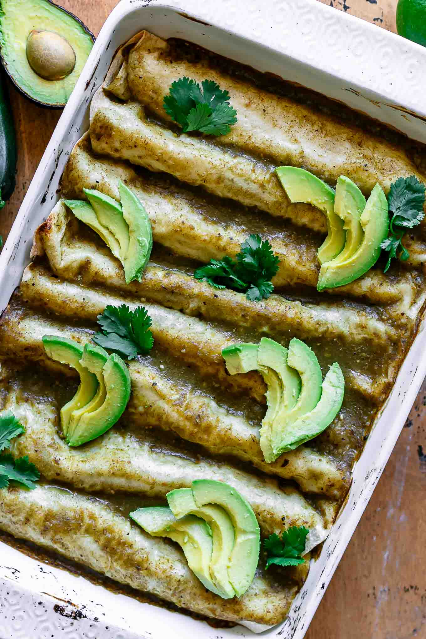 a white dish with zucchini enchiladas with green sauce topped with avocados and cilantro