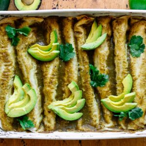 zucchini enchiladas with green enchilada sauce in a white baking dish and garnished with avocado slices and cilantro