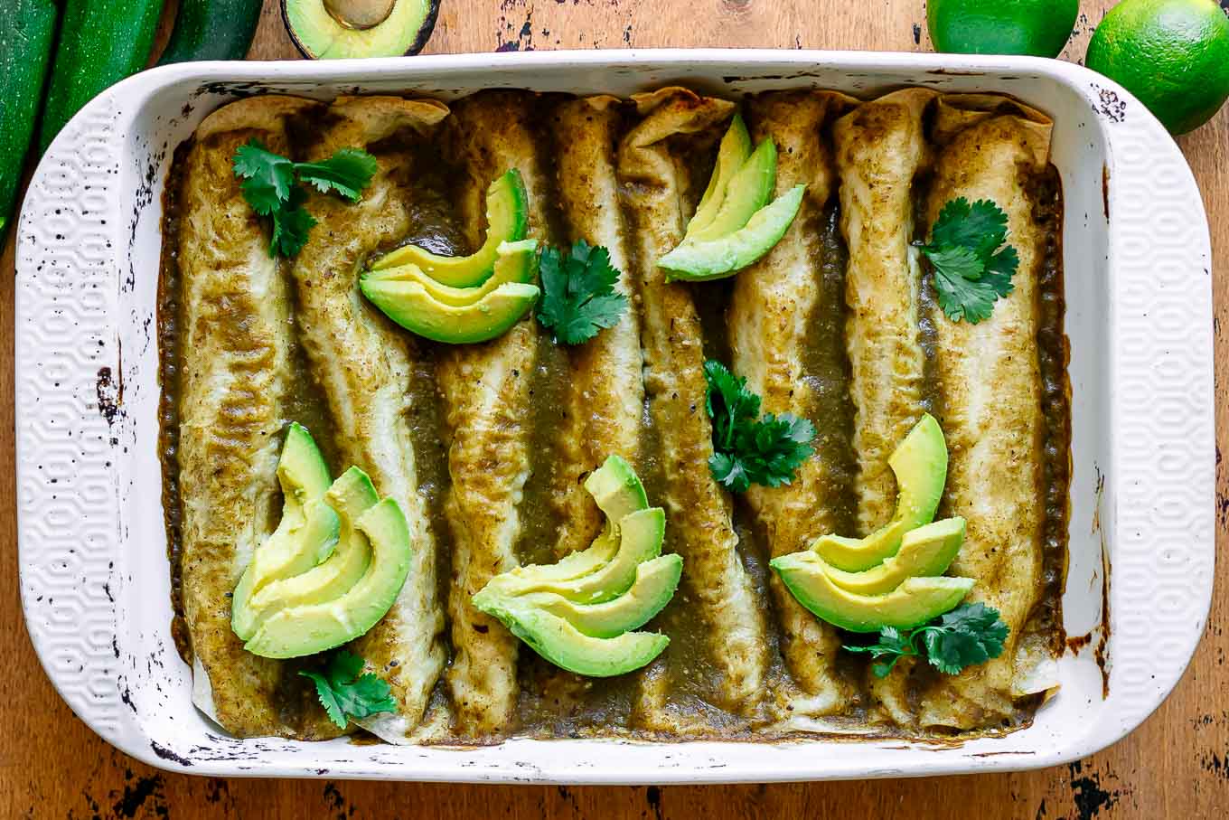 zucchini enchiladas with green enchilada sauce in a white baking dish and garnished with avocado slices and cilantro