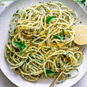 a white plate with lemon pesto pasta on a table with fresh lemons and fresh basil