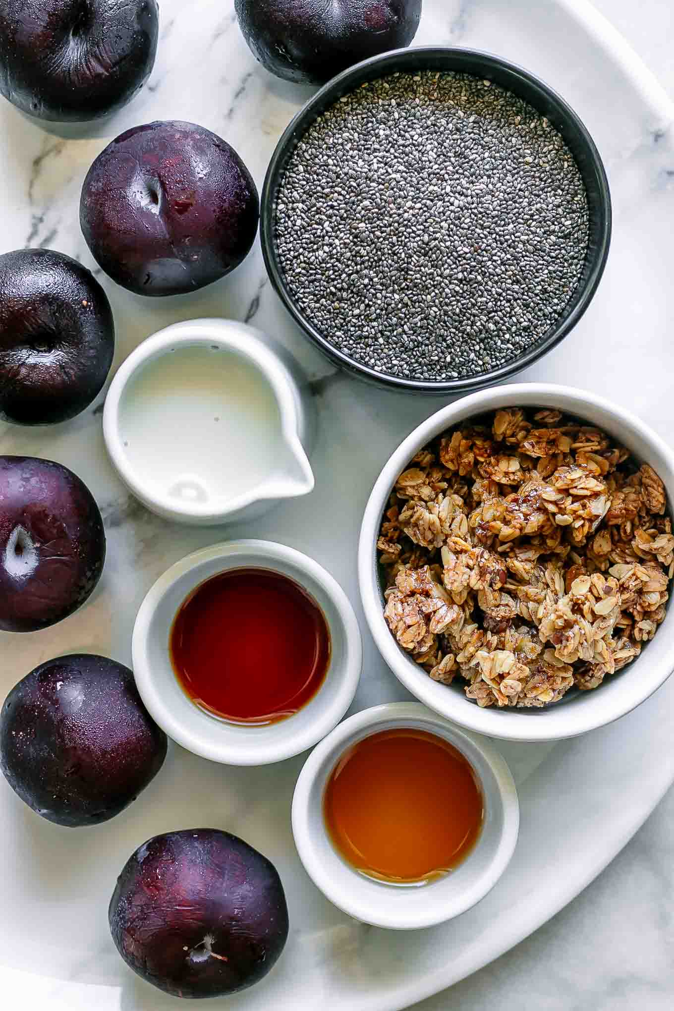 small bowls of chia seeds, plums, milk, vanilla, maple syrup, and granola on a white table