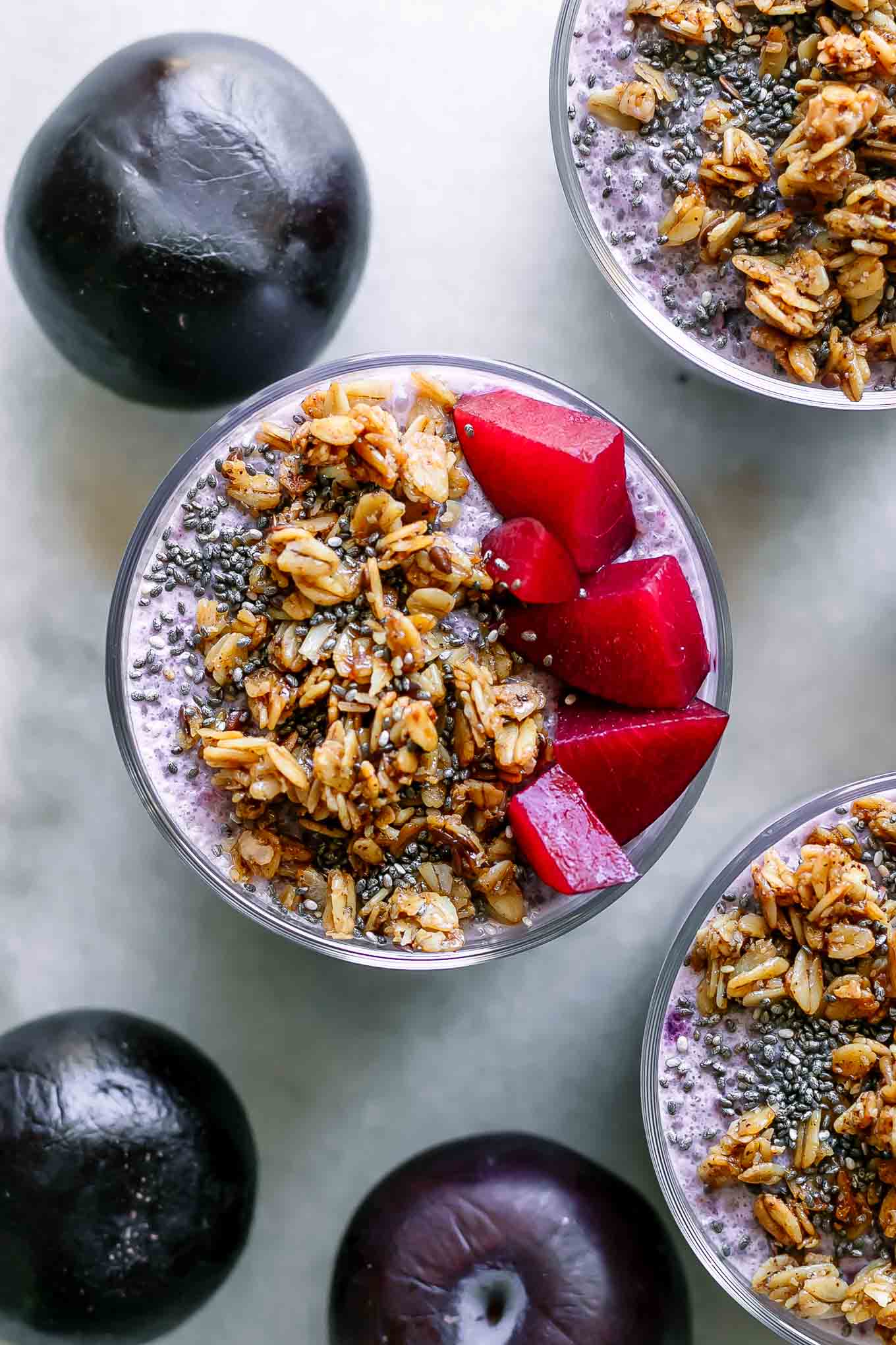 three small bowls of plum chia pudding with granola on a white table with purple plums