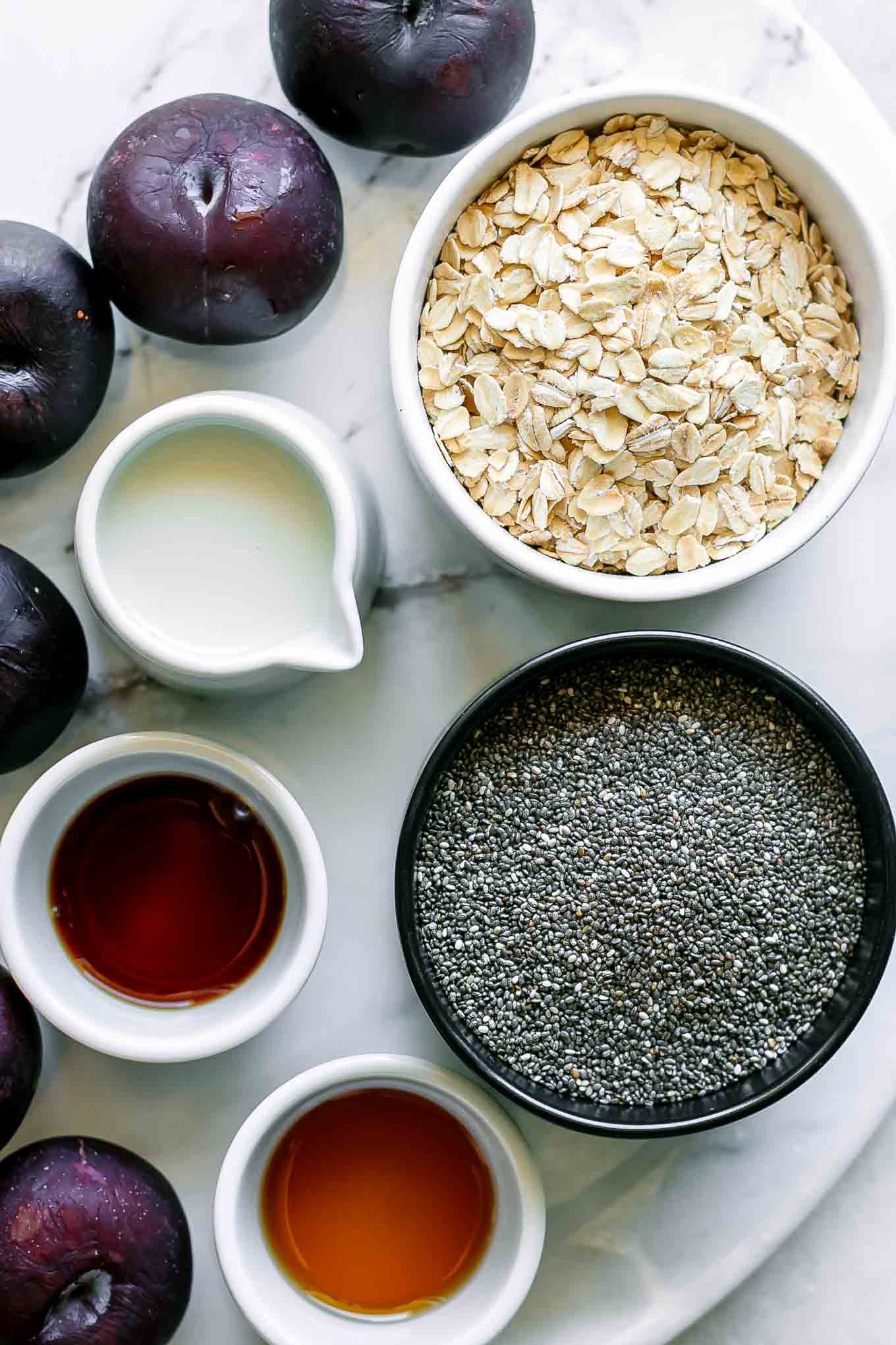 bowls of rolled oats, chia seeds, milk, vanilla, and maple syrup on a table with plums