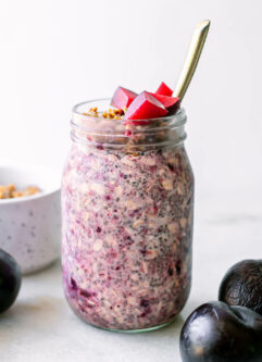 purple overnight oats with plums on top in a mason jar with a gold fork on a white table