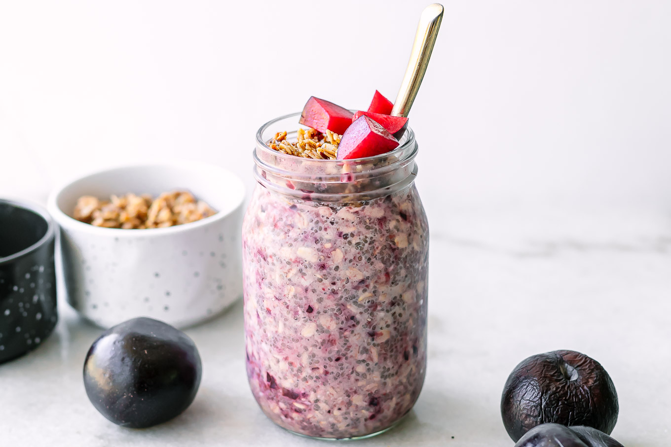 purple plum overnight oats in a jar with granola and cut plums as garnish on a white table