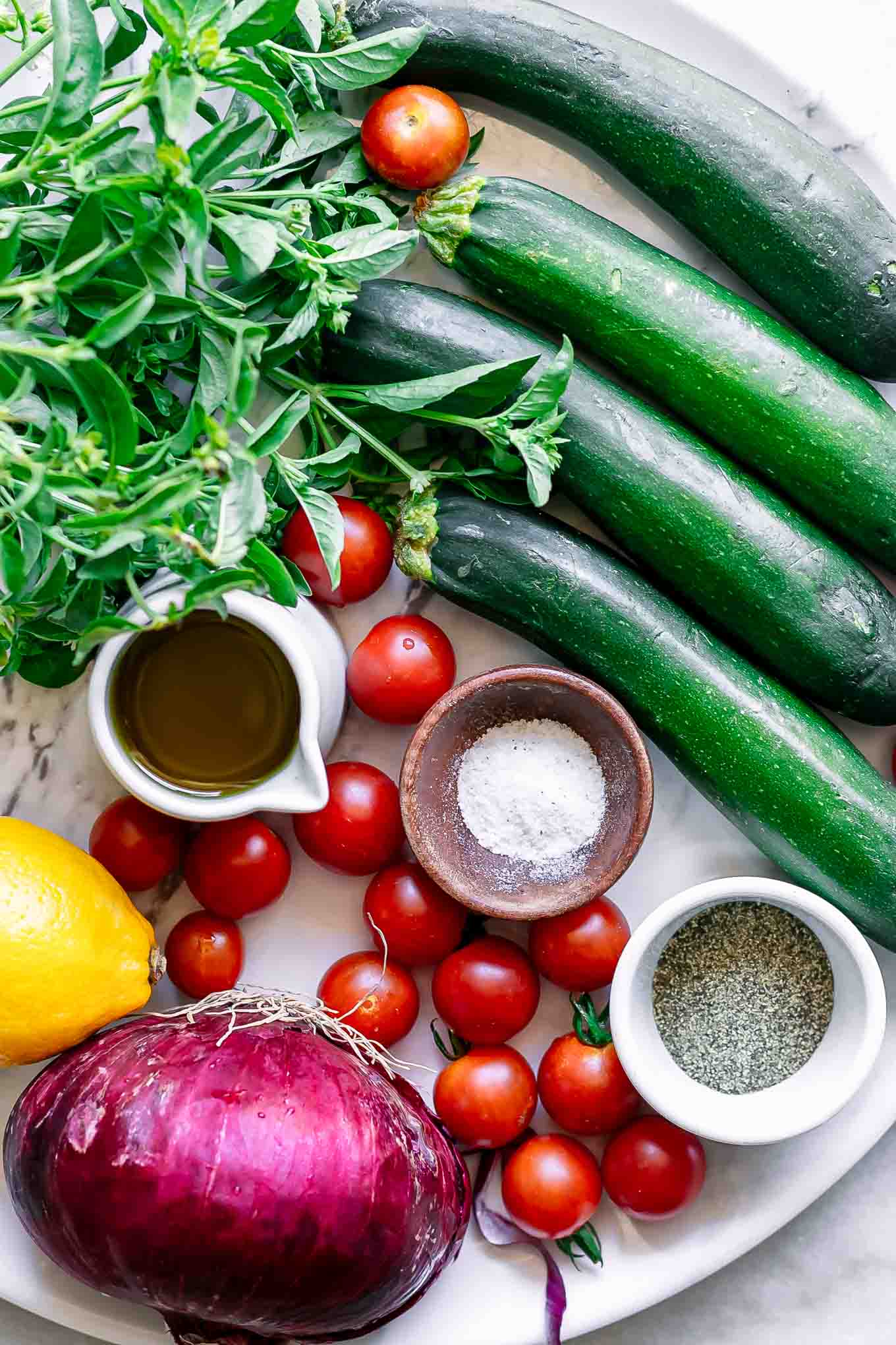 zucchini, tomatoes, fresh basil, red onion, lemon, and bowls of oil, salt, and pepper on a white table