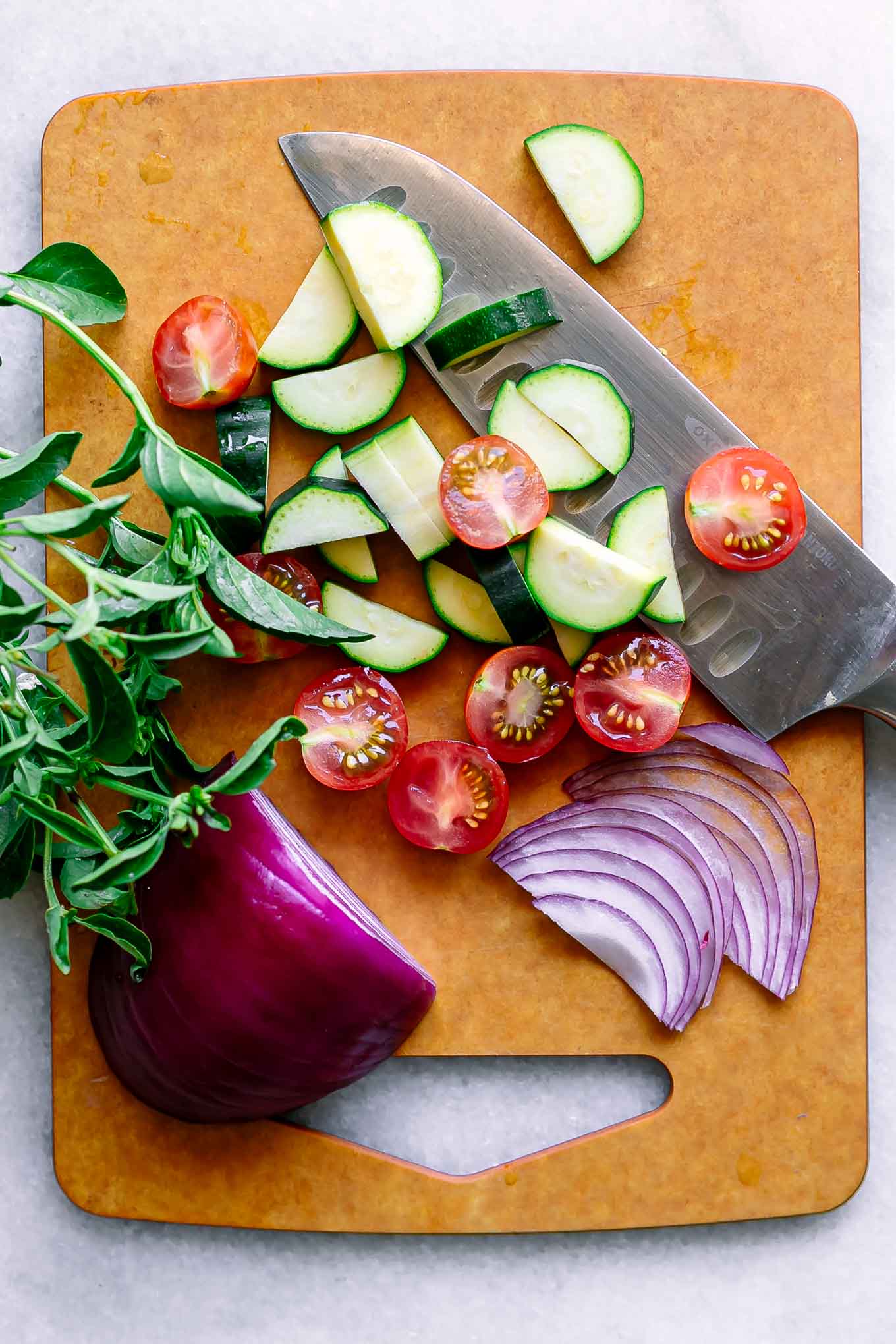 cut zucchini, tomatoes, and red onion on a wood cutting board with a knife