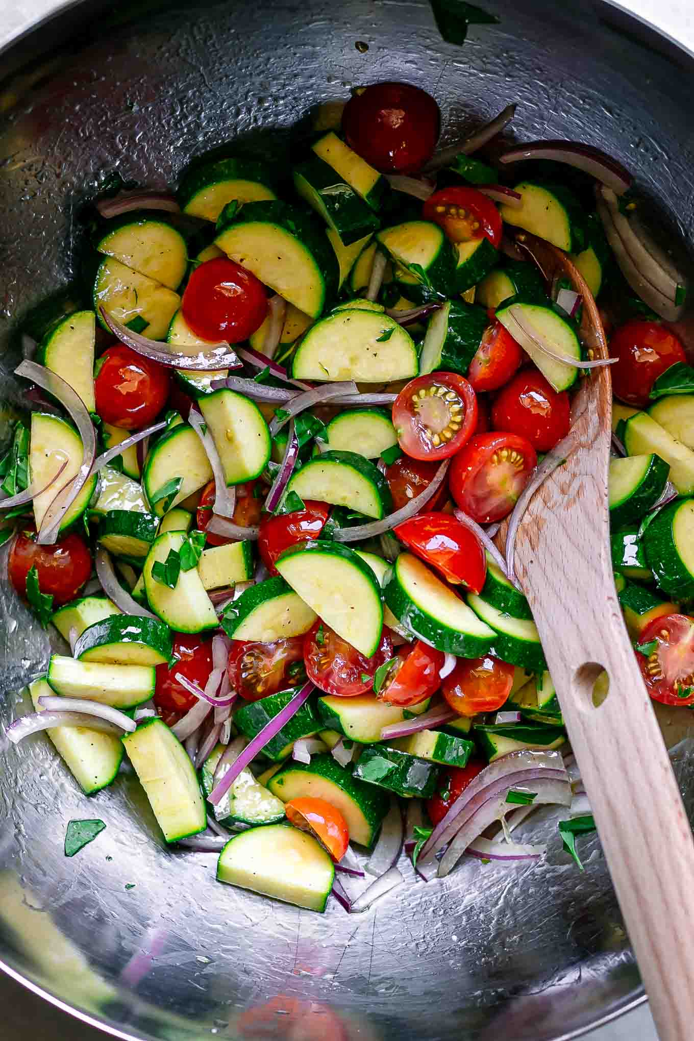 cut zucchini, tomatoes, onion, and basil inside a bowl with a wooden spoon