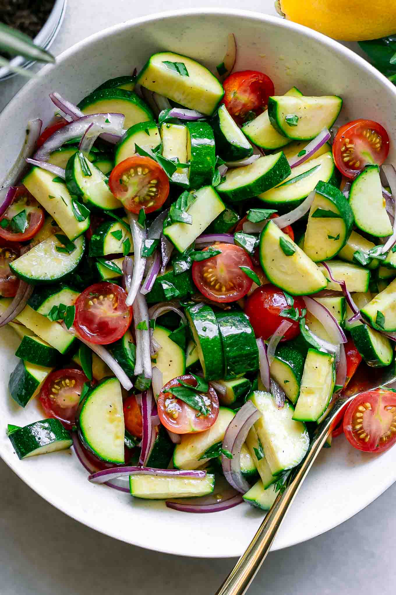 a close up photo of a raw zucchini and tomato salad in a white bowl with basil