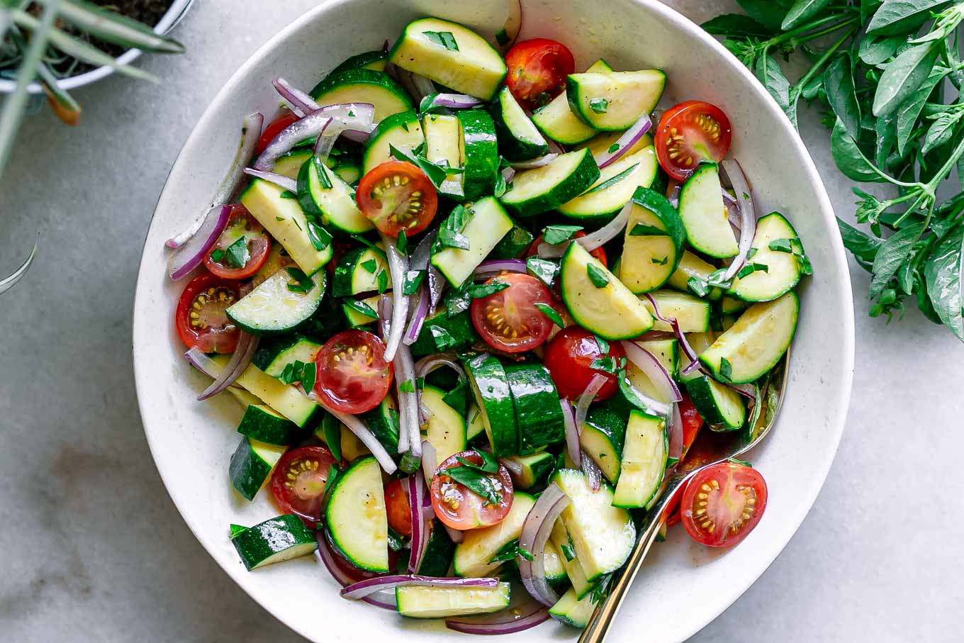 a raw zucchini tomato onion and basil salad in a white bowl on a white table with fresh basil