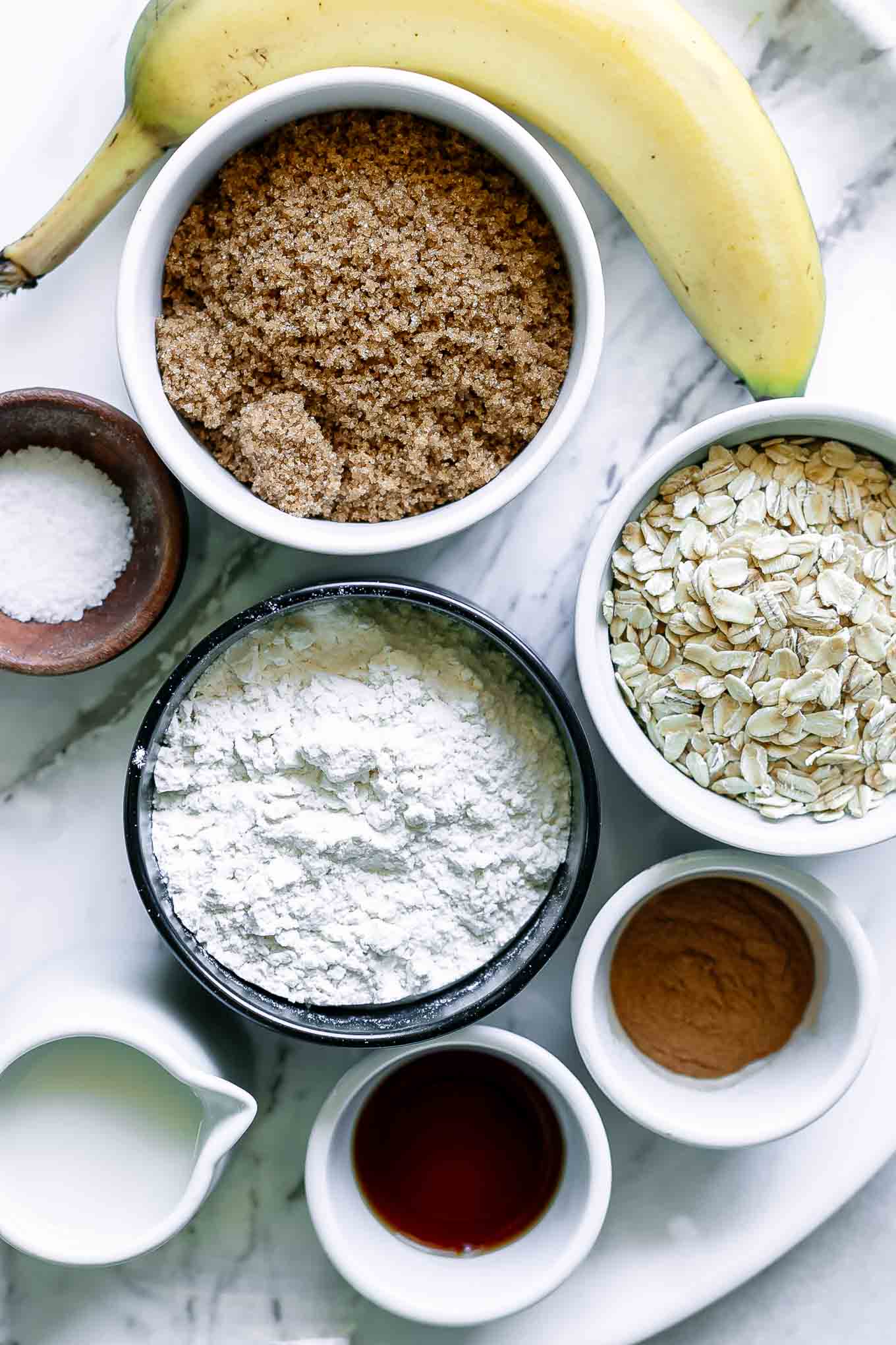 a banana with bowls of rolled oats, flour, brown sugar, and other ingredients for cookies