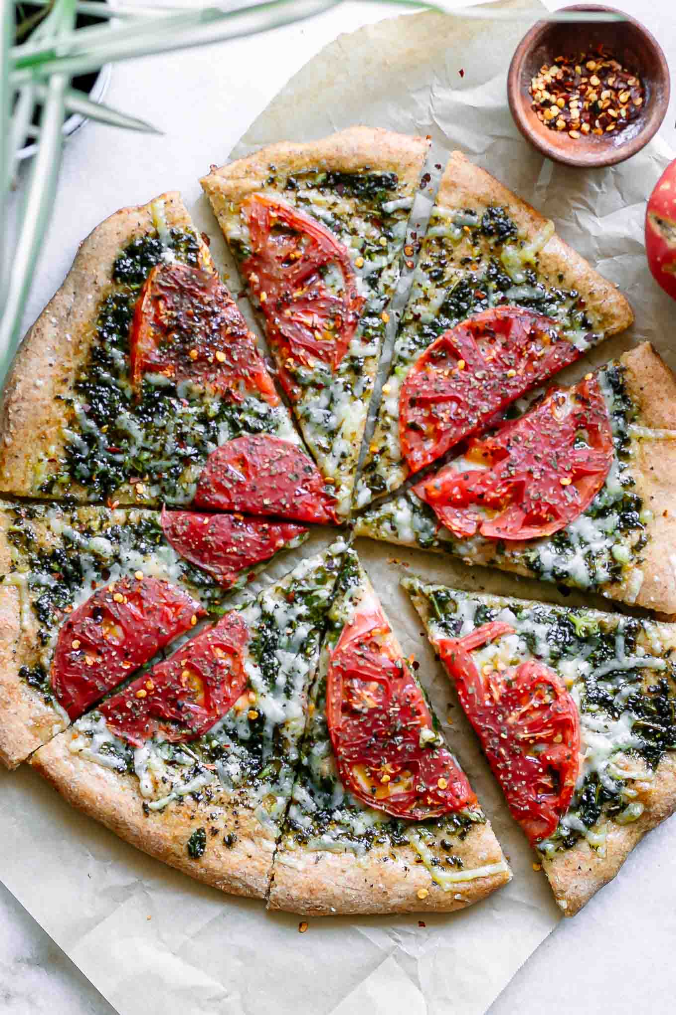 a vegan pizza with pesto sauce and sliced tomatoes on a white table with a bowl of red pepper flakes