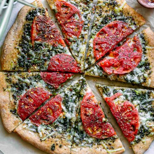 a sliced pesto pizza with roasted tomatoes on a white table with a tomato and a bowl of red pepper