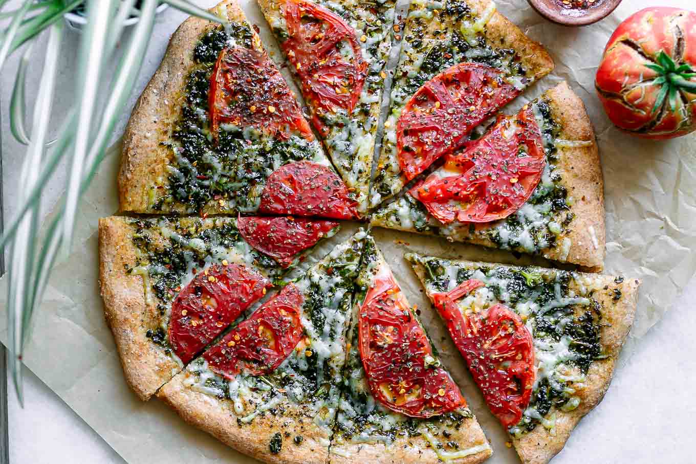 a sliced pesto pizza with roasted tomatoes on a white table with a tomato and a bowl of red pepper