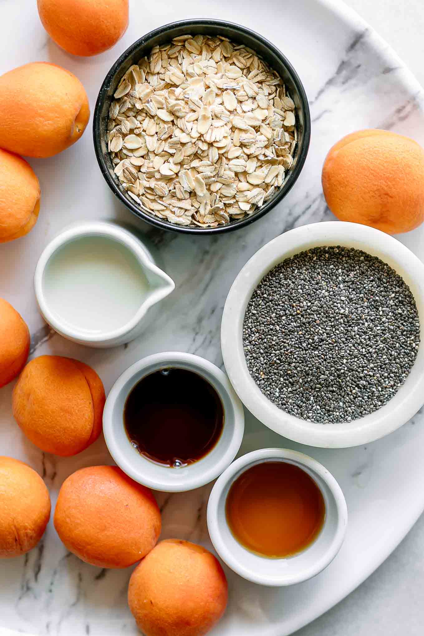 bowls of oats, chia seeds, milk, vanilla, syrup, and apricots on a white table