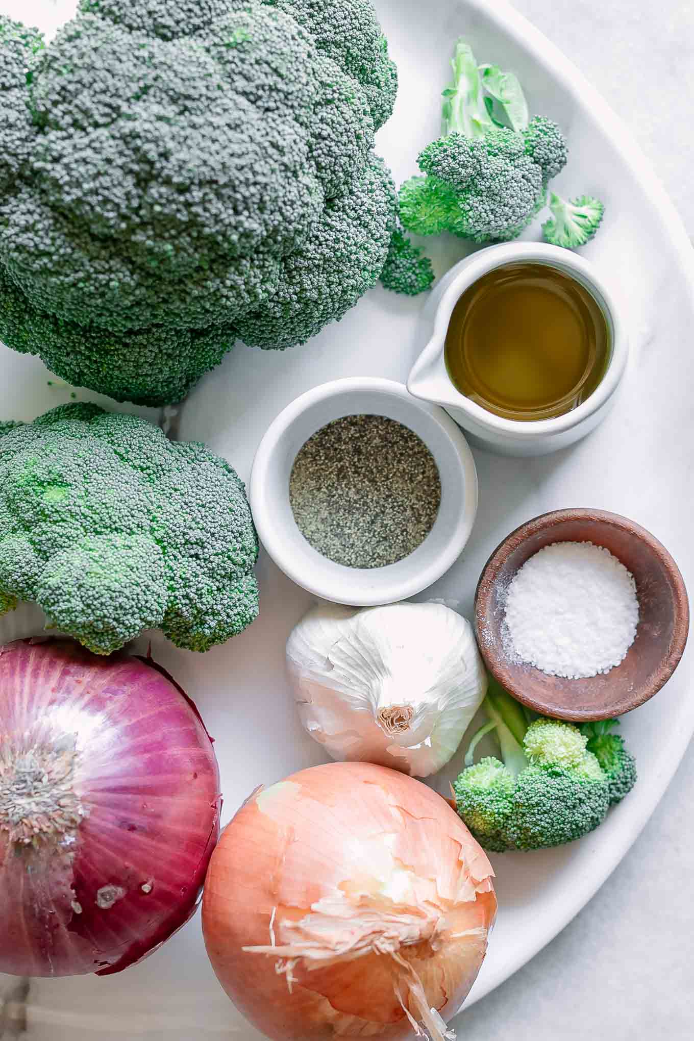 broccoli, onions, and bowls of oil, salt, pepper, and garlic on a white table before roasting