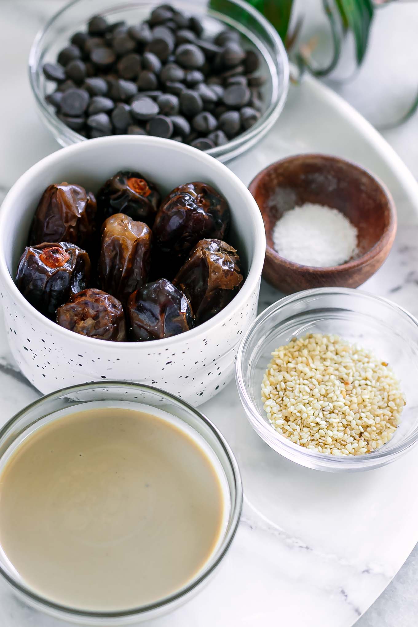 bowls of dates, tahini, chocolate chips, sesame seeds, and salt on a white table