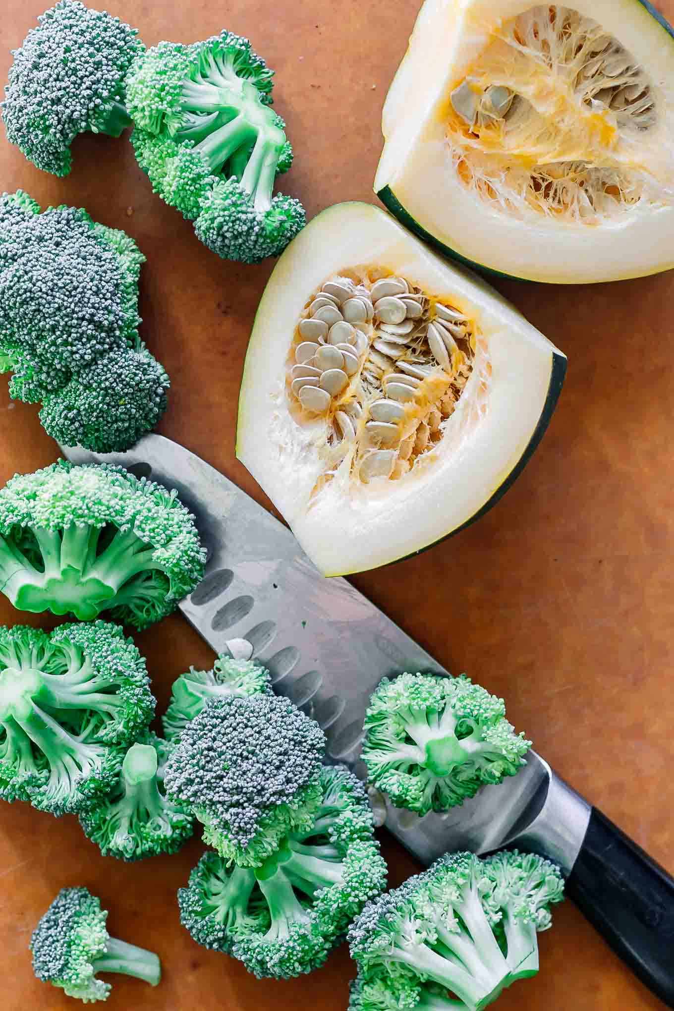 cut broccoli florets and sliced acorn squash on a wood cutting board with a knife