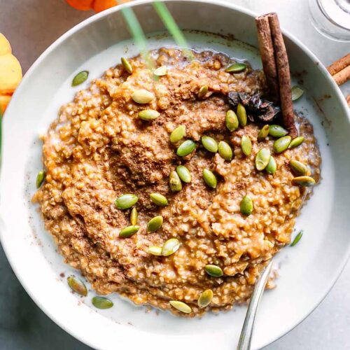 a bowl of pumpkin oatmeal with pumpkin seeds as a garnish and a cinnamon stick on a white table with a mini pumpkin