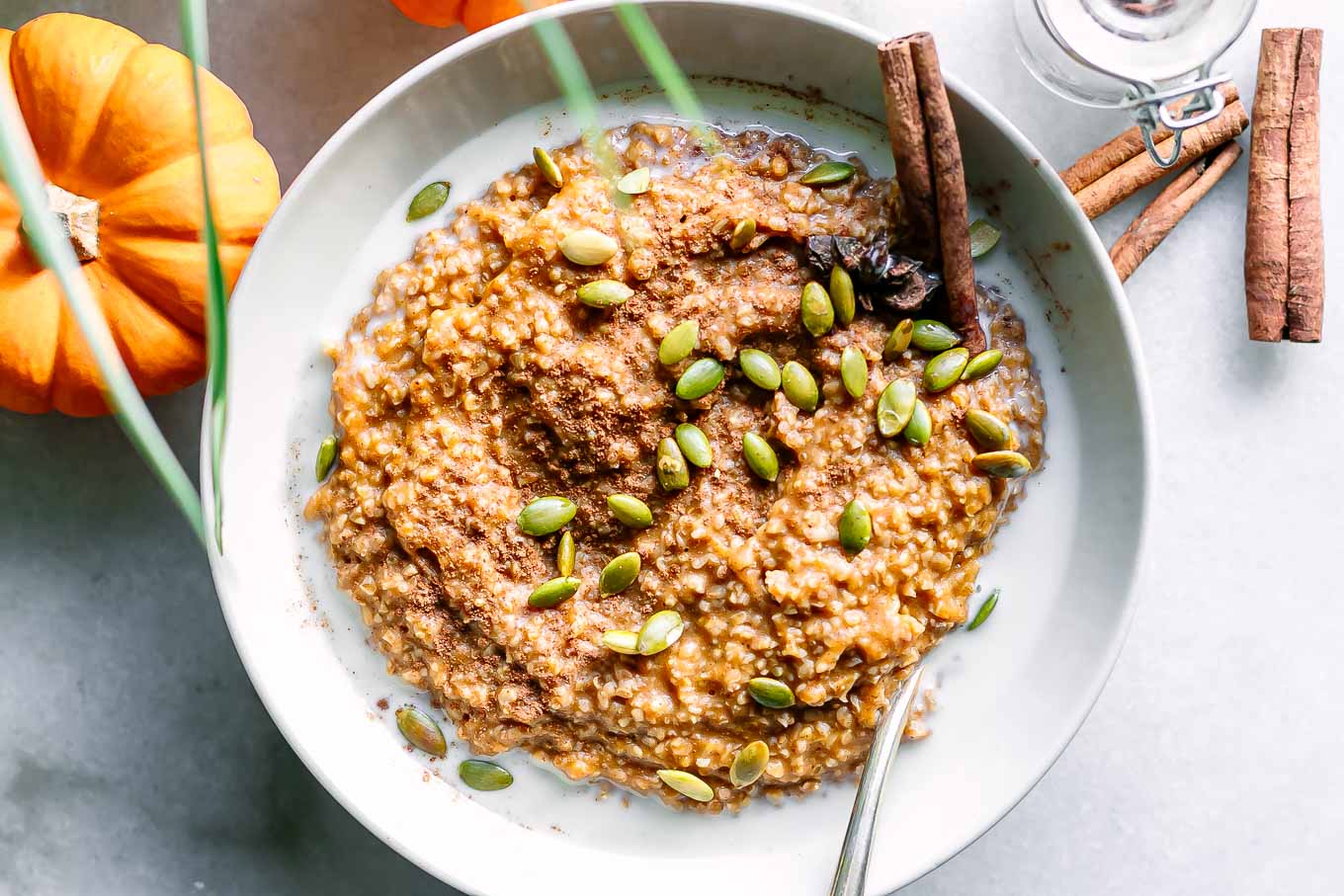 a bowl of pumpkin oatmeal with pumpkin seeds as a garnish and a cinnamon stick on a white table with a mini pumpkin