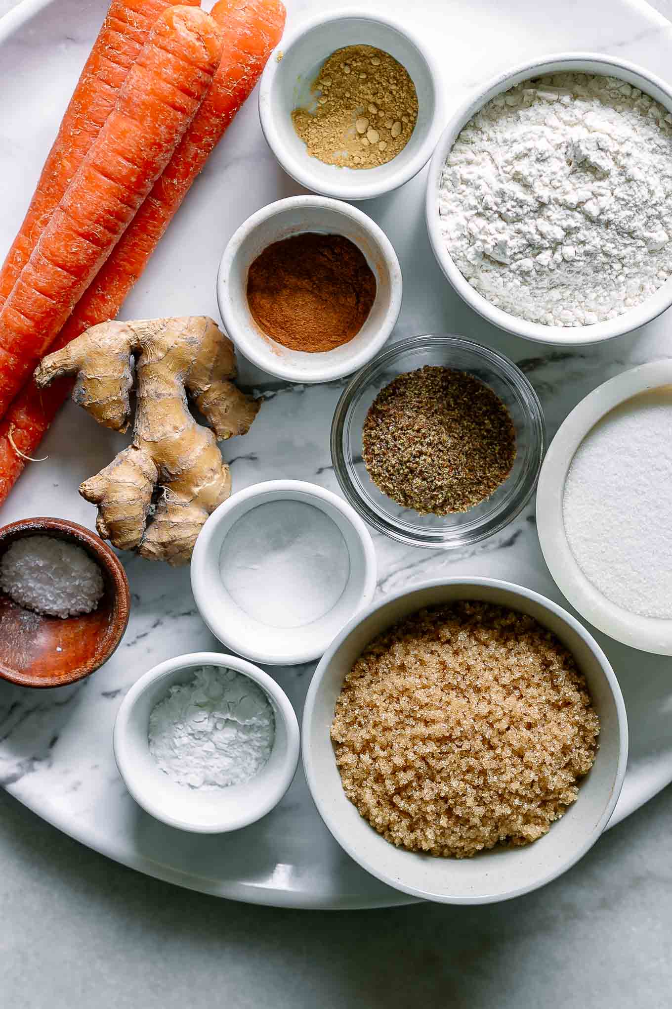 bowls of flour, sugar, vanilla, spices, and other ingredients for carrot cake muffins on a white table