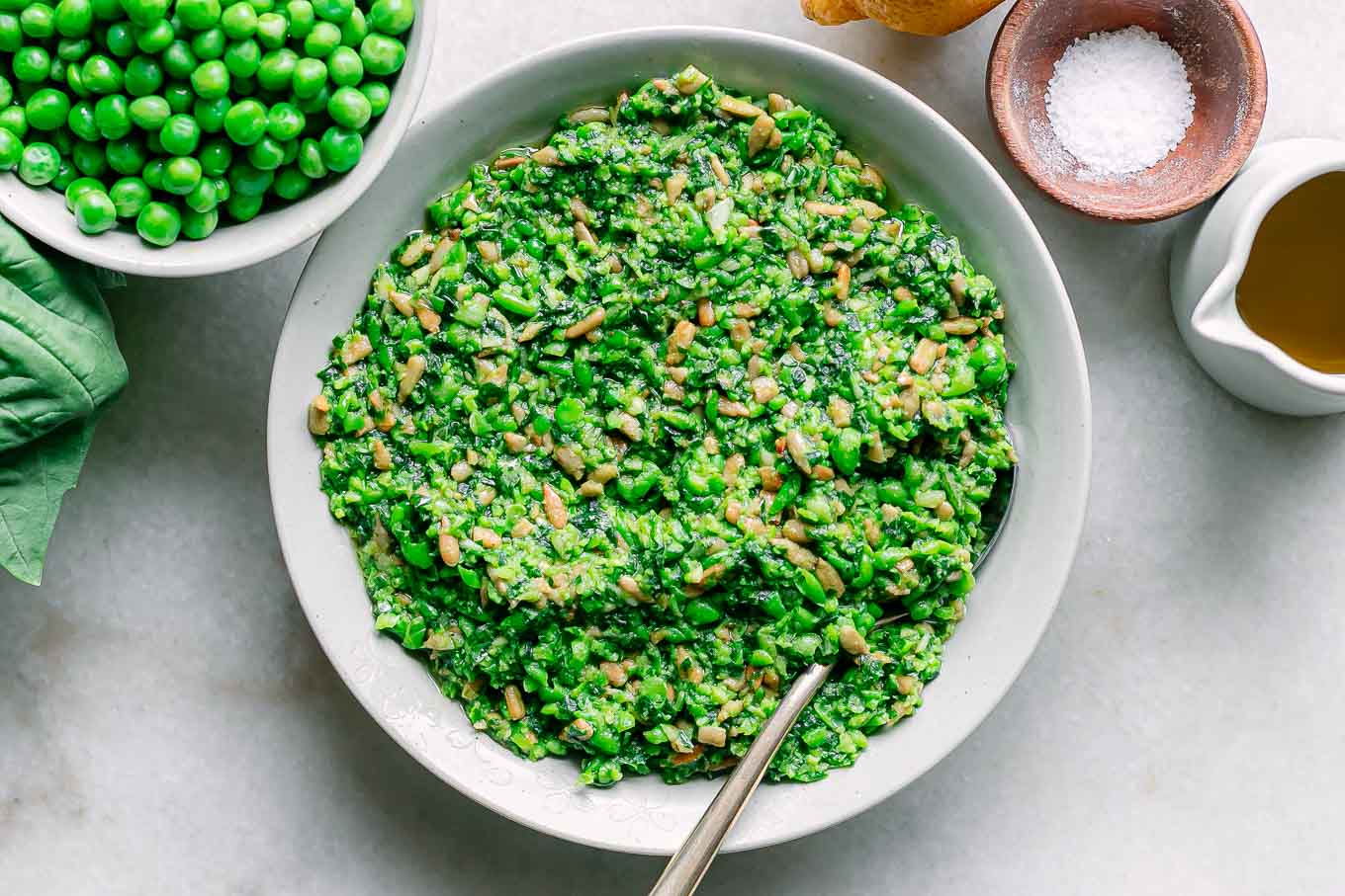 a bowl of green pea pesto on a white table with a bowl of fresh spring peas