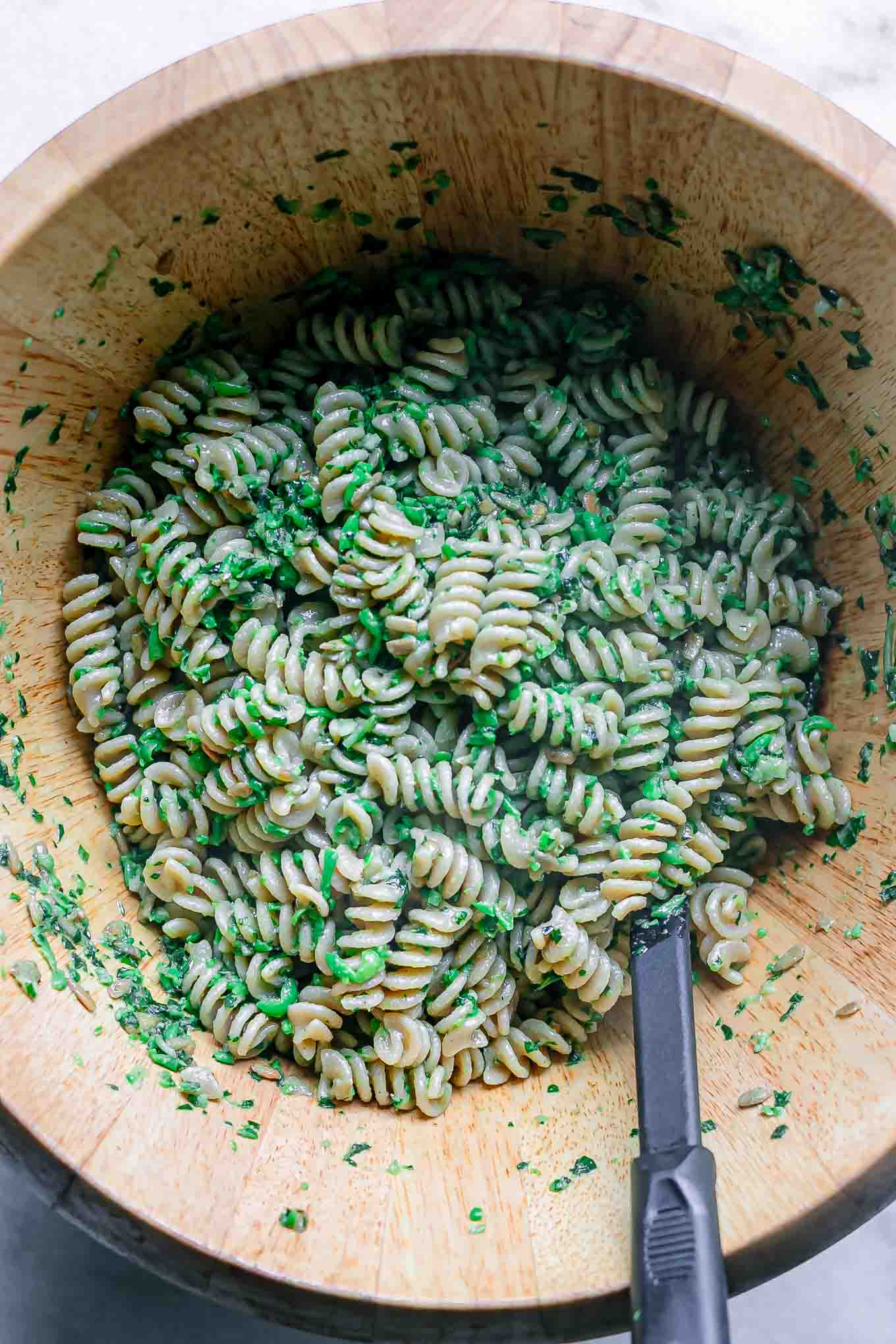 a large wooden mixing bowl with pasta and pesto sauce inside with a black spoon