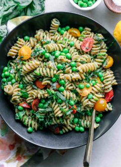 a bowl of pesto pasta salad with green peas, tomatoes, and basil on a white table with a gold fork