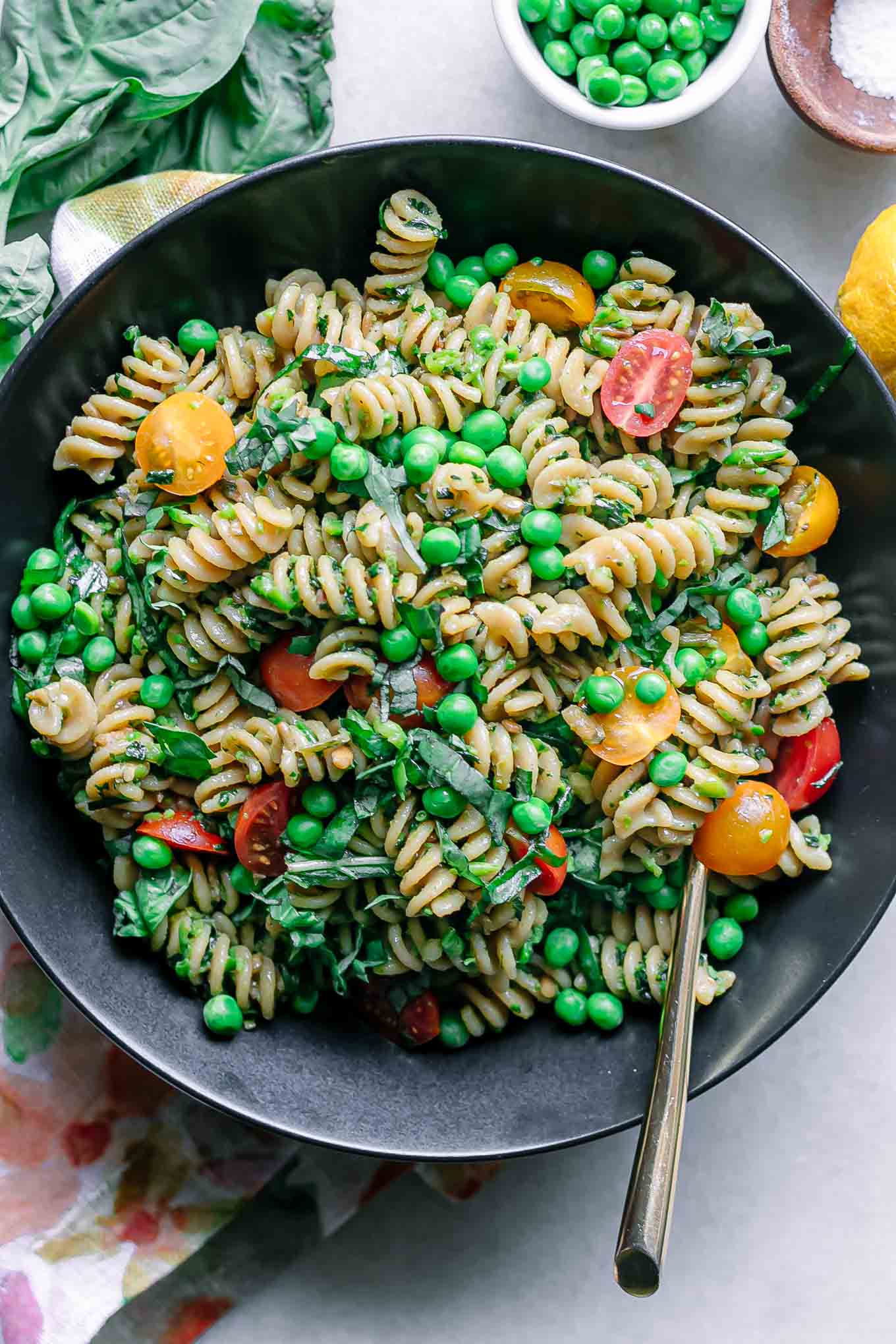 a bowl of pesto pasta salad with green peas, tomatoes, and basil on a white table with a gold fork