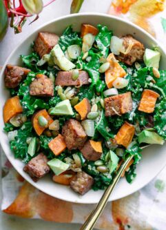 a white bowl with kale and quinoa salad with sweet potatoes and tempeh with avocado on a white table