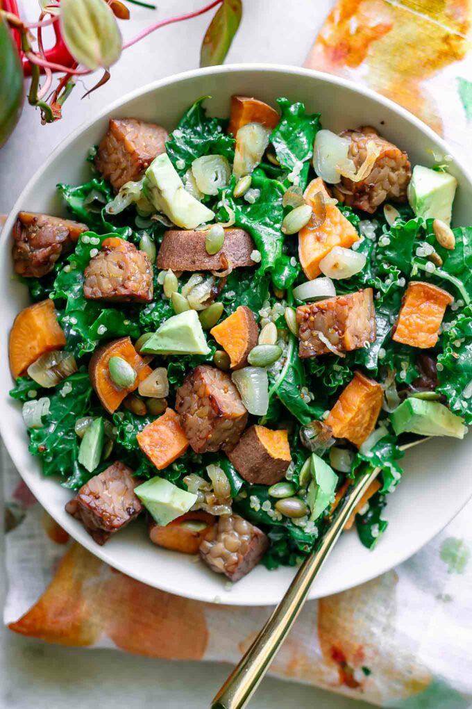 a white bowl with kale and quinoa salad with sweet potatoes and tempeh with avocado on a white table