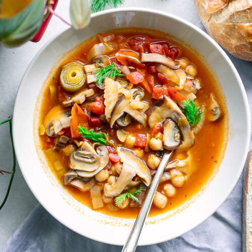 a white bowl of vegan cioppino soup with mushrooms on a table with sourdough bread for dipping