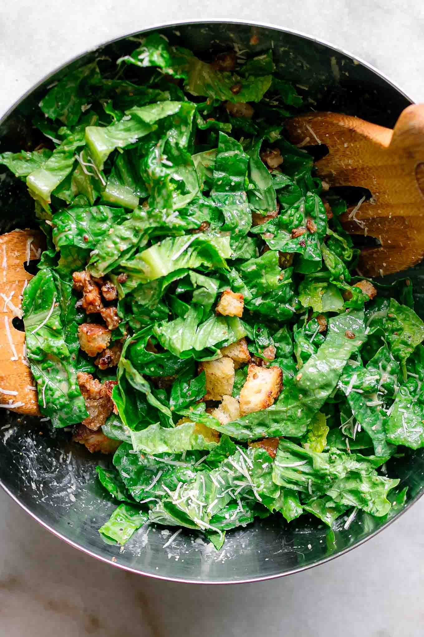 a mixing bowl with caesar salad ingredients inside with two wood tongs